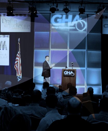 Speaker addressing a large audience at a conference with GHA branding.